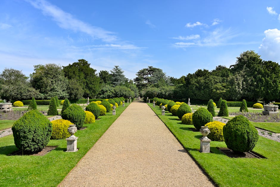 A traditional brick house with a pitched roof and multiple brick chimneys sits amidst a vibrant garden. The garden features a variety of flowering plants, including pink tulips and dark pink primroses