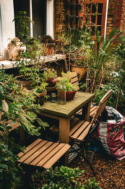 A cozy urban garden patio situated adjacent to a red brick building with a large window and a wooden door with glass panels. The space features a weathered wooden table surrounded by matching wooden c